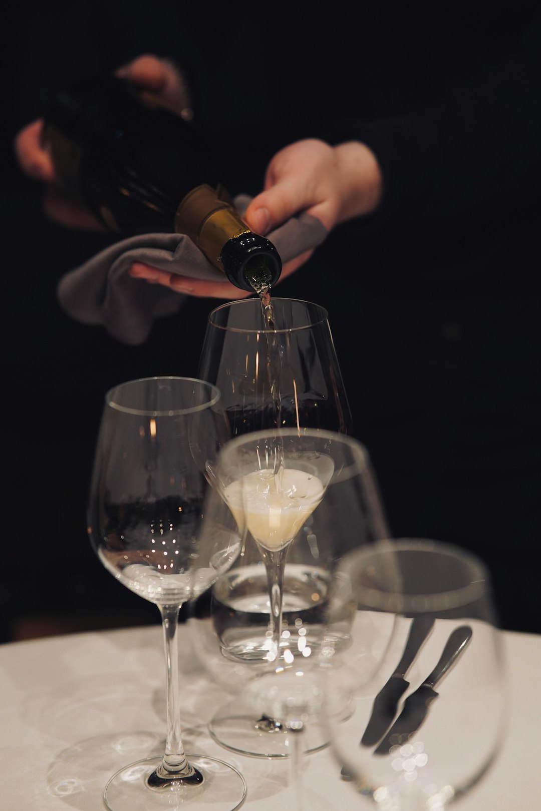 Ein Kellner schenkt Schaumwein in ein Glas auf weiß gedecktem Tisch ein, umgeben von leeren Weingläsern und Besteck.