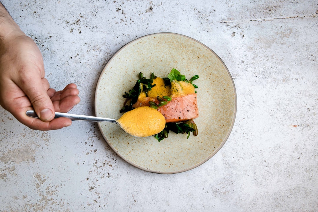 Gourmet-Teller mit Lachs, grünem Gemüse und orangefarbenem Schaum, angerichtet mit einem Löffel auf hellem Steintisch.