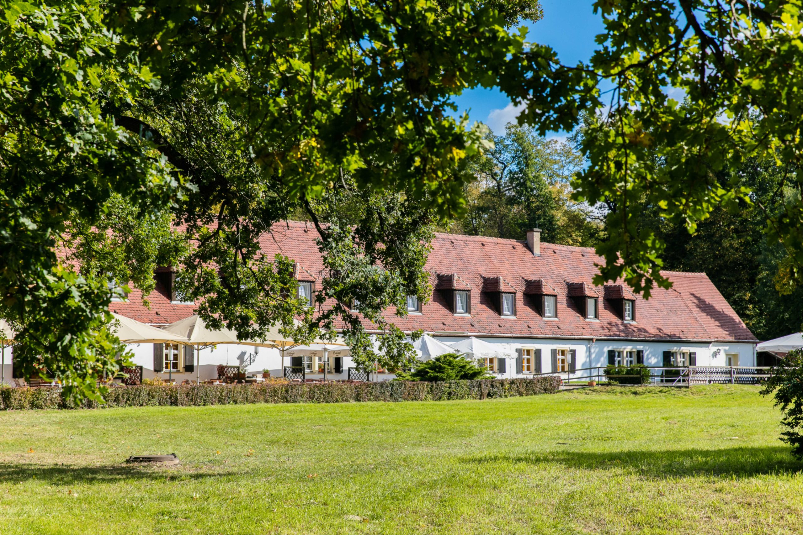 Historisches Hotel in Moritzburg mit rotem Dach und Sommerterrasse, eingebettet in grüne Wiesen und von alten Bäumen umgeben.