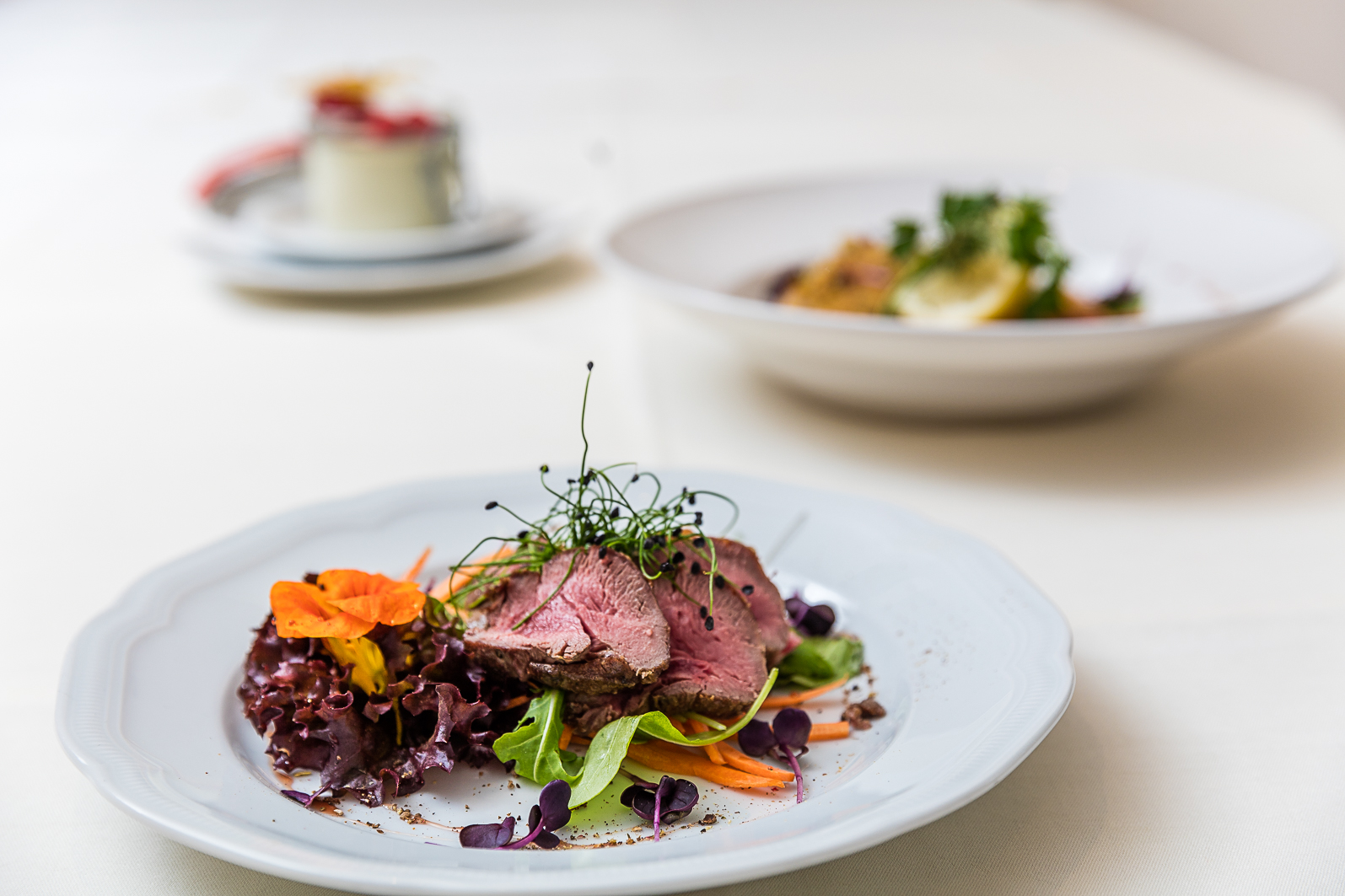 Gourmetteller mit rosa gebratenem Fleisch, frischem Gemüse und Kräutern, elegant serviert auf weiß gedecktem Tisch.