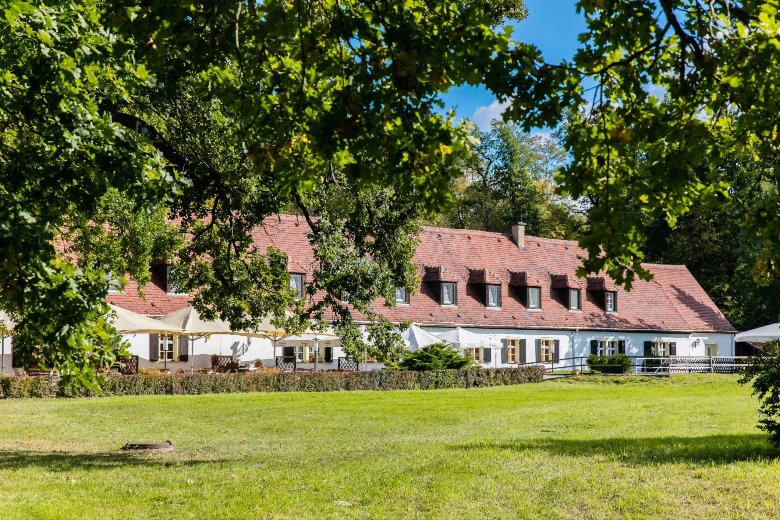 Historisches Hotel in Moritzburg mit rotem Dach und Sommerterrasse, eingebettet in grüne Wiesen und von alten Bäumen umgeben.
