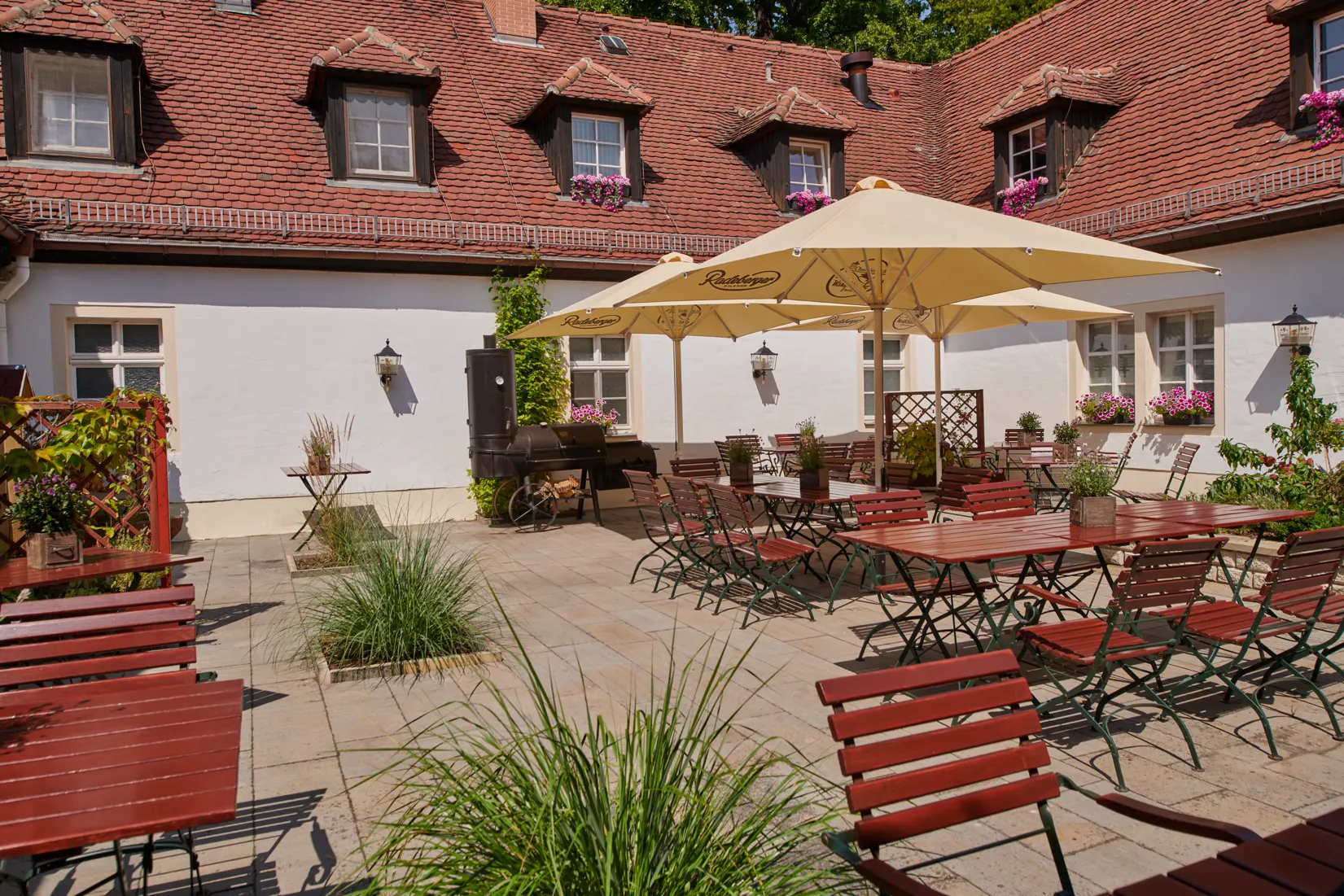 Innenhof – Außengastronomie der Churfürstlichen Waldschänke Moritzburg Inner courtyard of the Churfürstliche Waldschänke Moritzburg with beer garden seating, large parasols and smoker grill on the historic façade