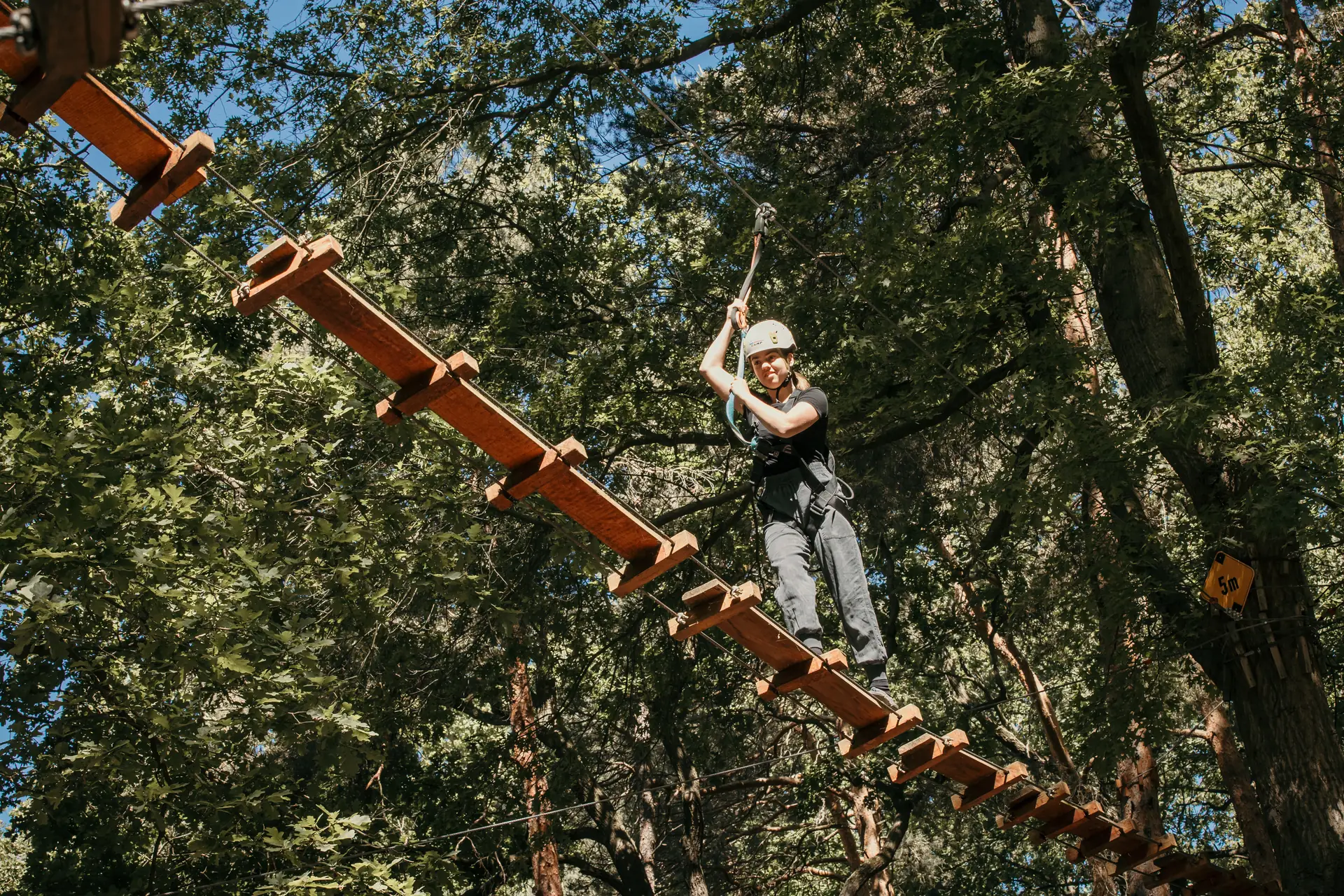 Frau mit Helm und Klettergurt steigt über eine schräge Holzleiter in den 10-Meter-Parcours des Hochseilgartens Aufstieg in den 10-Meter-Parcours