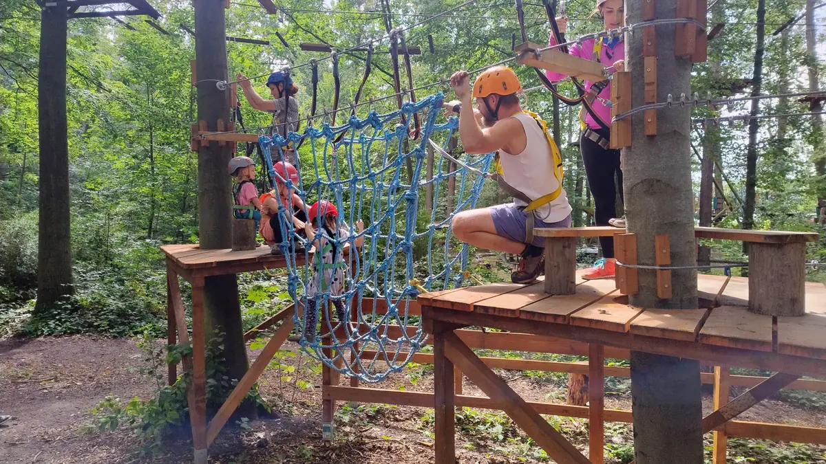 Kinderklettern im Niedrigseilgarten Kinder mit Helm und Klettergurt überqueren ein blaues Kletternetz im Niedrigseilgarten zwischen Holzplattformen