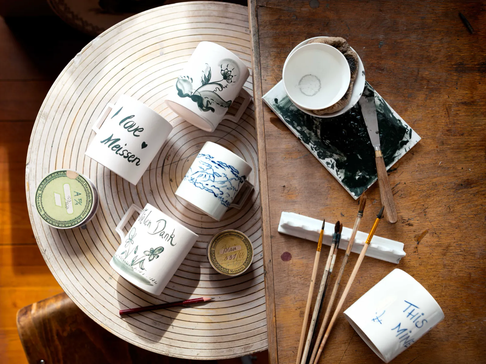 meissen_diy_tassen-mit-handschrift_2 Porzellantassen mit Aufschriften wie „I love Meissen“ und Blumenmotiven auf einer Drehscheibe, daneben Pinsel und Farbpalette, von oben fotografiert